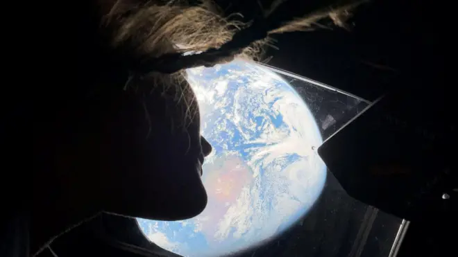 A imagem mostra a silhueta de uma astronalta, dentro da espaçonave Orion, observando a Terra através de uma janela. O rosto aparece em perfil, parcialmente iluminado pelo brilho intenso do planeta azul ao fundo, a Terra, destacando os contornos do cabelo que flutua levemente na ausência da gravidade. A Terra ocupa boa parte da cena, com nuvens brancas e oceanos visíveis, criando um contraste entre a escuridão do espaço e a luminosidade do planeta.