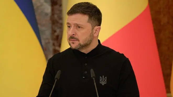 Volodymyr Zelensky yambaye umupira w'amaboko maremare umeze nk'ishati, ahagaze imbere y'ibendera ry'Ububiligi n'ibendera rya Ukraine. 