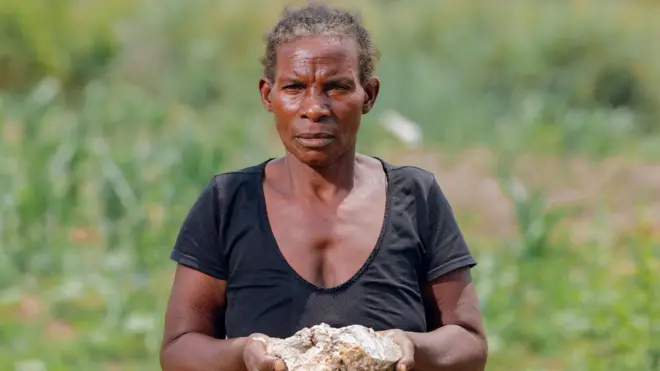 A photo portrait of Mary, a Zambian farmer. She stands in the centre of the frame looking pensive and holding up a large lump of white contaminated crop soil, with both hands. There is greenery behind her and she wear a black top and patterned skirt. 