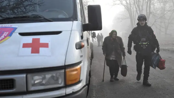 Поздняя осень, туман. По дороге идёт пожилая женщина с тростью, на ней пальто и платок, рядом с ней полицейский несёт её сумки. Слева от них на дороге стоит белый минивэн с красным крестом и эмблемой со словом «эвакуация».