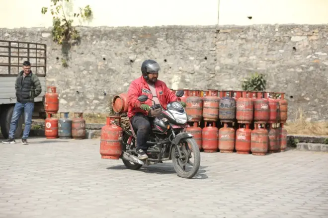दुईवटा ग्यास सिलिन्डर राखेर कालो रङ्गको मोटरसाइकल चलाइरहेका रातो ज्याकेट लगाएका एक व्यक्ति