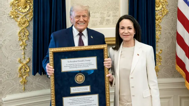 ダークスーツを着たトランプ氏が、ノーベル平和賞のメダルが入った金色のフレームを両手で持ち、笑顔でカメラを見ている。左隣りには白いスーツを着たマチャド氏が立ち、フレームを片手で持ちながらほほえんでいる