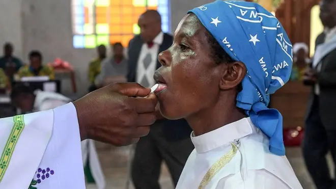 The white embroidered sleeve of a Catholic cleric in Kenya placing a communion wafer of the tongue of a kneeling worshipper wearing white robes and a blue headscarf