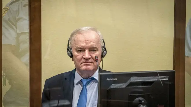 Ратко Младич в костюме с галстуком сидит за стеклом, на нем наушники, перед ним микрофон и монитор.