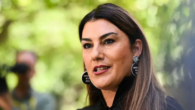 Senator Lidia Thorpe at a press conference. She has long brown hair, is wearing circle earrings and trees are out of focus in the background