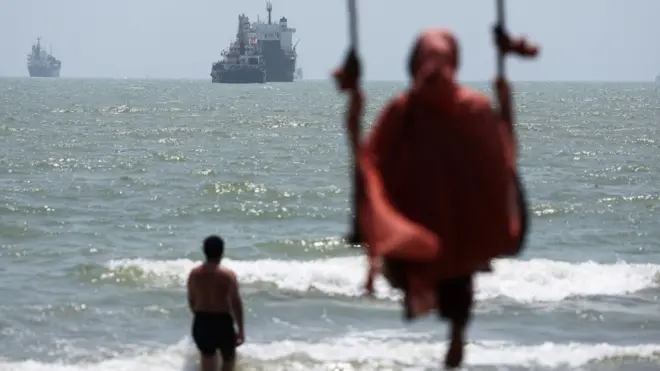 плажа у ирану, ормуски мореуз, поглед на ормуски мореуз са иранске обале, жена се љуља у љујашци, а мушкарац је у води и гледа у бродове у ормуском мореузу