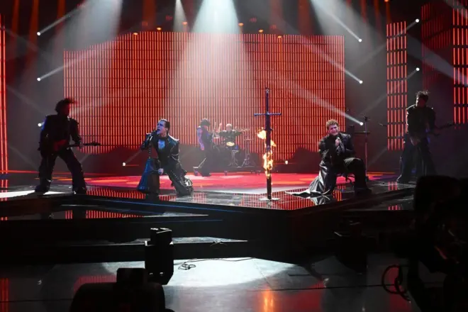 muškarci klečeći pevaju na sceni, obučeni su u crno, između njih je mač koji gori 