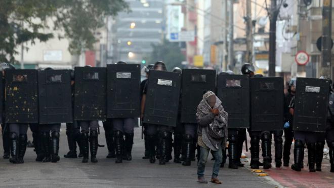 Homem se protegendo na frenteaposta de timebarreiraaposta de timepoliciais com escudos, na Cracolândia