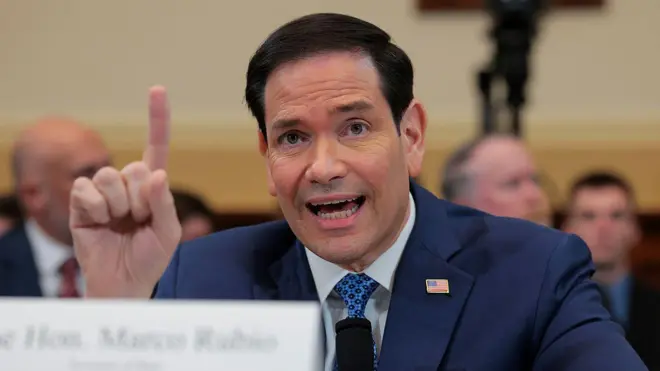 Minisitiri w’ububanyi n’amahanga w’Amerika Marco Rubio arimo gutanga ubuhamya muri komite ishinzwe ububanyi n’amahanga yo mu nteko ishingamategeko y’Amerika, umutwe w’abadepite, mu nyubako yitwa ‘Rayburn House Office Building’, i Washington DC, muri Amerika, ku itariki ya 3 Kamena (6) mu 2026. Uko kumwumva kwabaye mu rwego rwo gusuzuma ubusabe bwa minisiteri y’ububanyi n’amahanga bw’ingengo y’imari y’umwaka w’imari wa 2027. Rubio yambaye ikositimu y'ubururu bwijimye, ishati y'umweru, na karuvati irimo amabara y'ubururu n'umukara. Ikositimu ye iriho umudari muto w'ibendera ry'Amerika. Aricaye, arimo kuvugira mu ndangururamajwi y'umukara, atunze urutoko hejuru, inyuma ye hari abantu benshi bataboneka neza. 