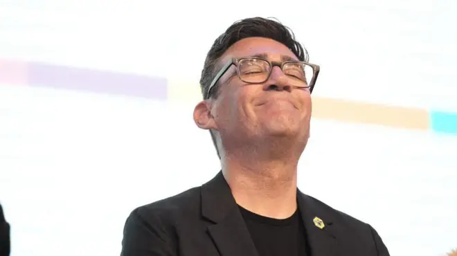 白い背景の檀上に黒い上着と丸襟のシャツ姿で立った、眼鏡をかけたバーナム氏が、両目を閉じて口をぎゅっと結んで微笑み、上を向いている