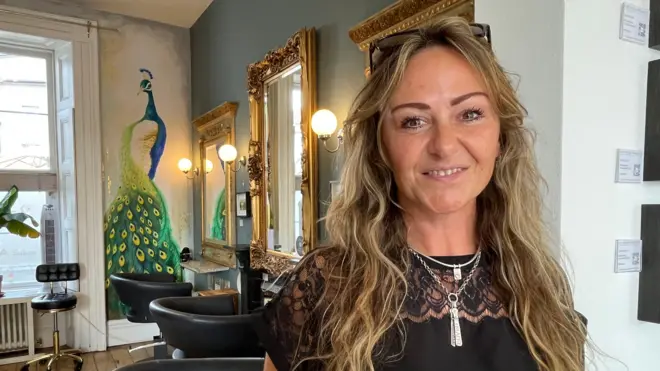 En el interior de una peluquería, una mujer de cabello rubio y ondulado sonríe directamente a la cámara, con los ojos brillantes. Al fondo, un cuadro de un pavo real adorna una pared, junto a grandes espejos con marcos dorados y sillones negros para los clientes.