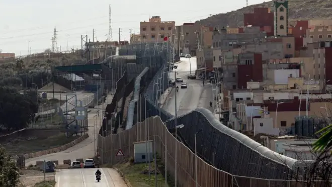 صورة التُقطت من الجانب الإسباني في 24 فبراير/شباط 2024 تُظهر الجدار والسياج اللذين يُحددان الحدود بين المغرب (يمين) وجيب مليلية الإسباني في شمال أفريقيا (يسار)