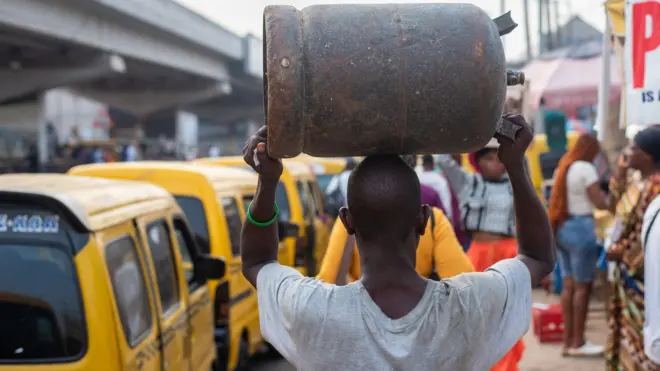 Ọkùnrin kan gbé agolo gáàsì ìdáná sórí, àwọn ọkọ̀ àti èrò wà ní ibi tó wà