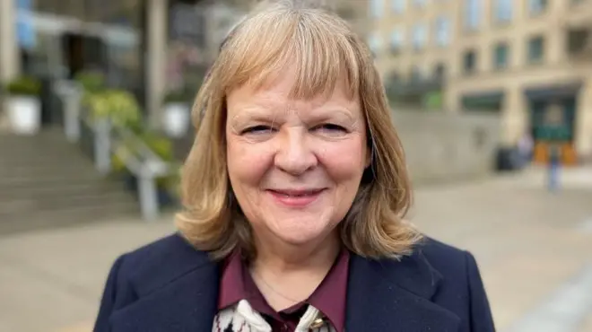 Una mujer que sonríe a la cámara, tiene el pelo rubio y lleva una chaqueta azul oscuro sobre una blusa burdeos y blanca