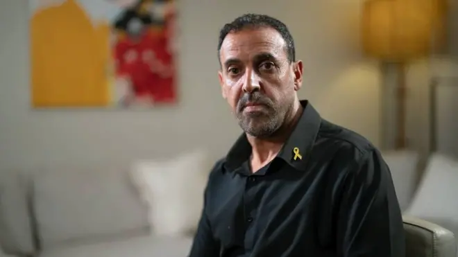 Portrait of Eli Sharabi in a black shirt with a yellow ribbon lapel pin, sitting in a room with largely neutral colours