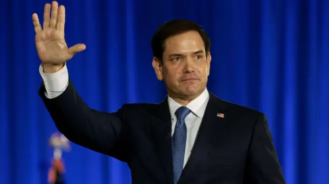 Marco Rubio com a mão para cima, acenando para pessoas
