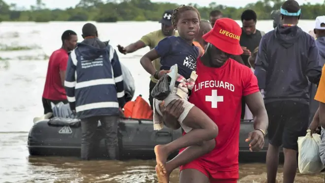 Um homem com uma camiseta vermelha e um chapéu com a palavra “Salva-vidas” ajuda uma menina a atravessar as águas da enchente no sul de Moçambique