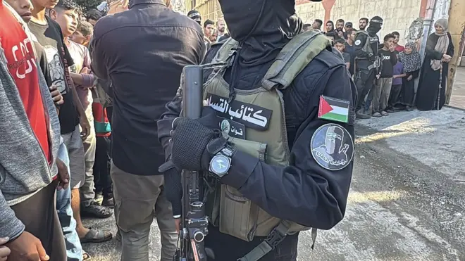 مقاتلون من حركة حماس يقفون استعداداً لتسليم الرهائن الإسرائيليين العشرين إلى فرق اللجنة الدولية للصليب الأحمر، ويظهر في الصورة مقاتل وجهه ملثم ويرتدي درعاً واقياً كُتب عليها "كتائب القسام"، وأعلى ذراعة شارة عليها وجه ملثم وكتب عليها "وحدة الظل"، بينما يحمل سلاحاً نارياً.