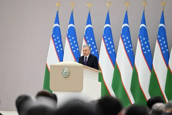 Shavkat Mirziyoev minbarda nutq so'zlamoqda. orqasida O'zbekiston bayroqlari.