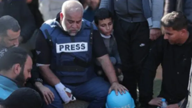 الصحفي الدحدوح مرتدياً سترة الصحفيين، يده مصابة ويجلس حزيناً بين جمع من الناس.