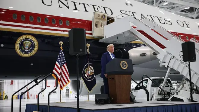 Trump standing at a podium on a stage in front of the new jet, painted white with dark red and blue stripes. There is a presidential seal and the words "United States of" visible.