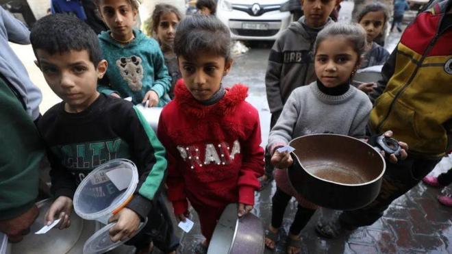 Crianças com potes e panelas vazias esperam enquanto profissionais de ajuda humanitária distribuem alimentos em Gaza no início deste mês