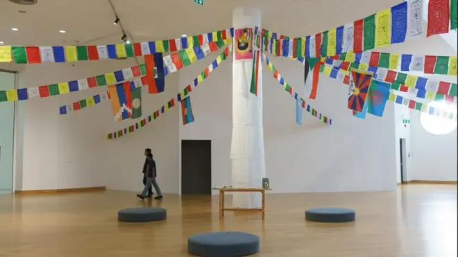 A large pillar stands in the middle of a gallery wrapped in white cloth, with several strings of colourful bunting carrying the flags of various countries and Tibetan art tied to the top of the pillar where a red and yellow flag has been pinned. There is a small wooden table at the base of the pillar as well as black circular cushions placed around it. Two people dressed in jeans and dark T-shirts are walking across the gallery