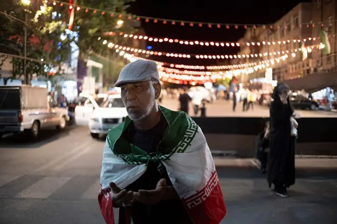 مؤيد للحكومة الإيرانية أسمر البشرة لديه لحية خفيفة بيضاء اللون، يرتدي العلم الإيراني يرفع يديه داعياً.