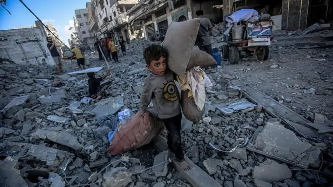 Израил Газадагы Абу Хасира көчөсүнө чабуул койгондон кийин палестиналык жаш балдар жарактуу буюмдарын терип жатышат. Арт жакта бир нече имарат толугу менен кыйраганын көрүүгө болот. 2025-жылдын 30-сентябры