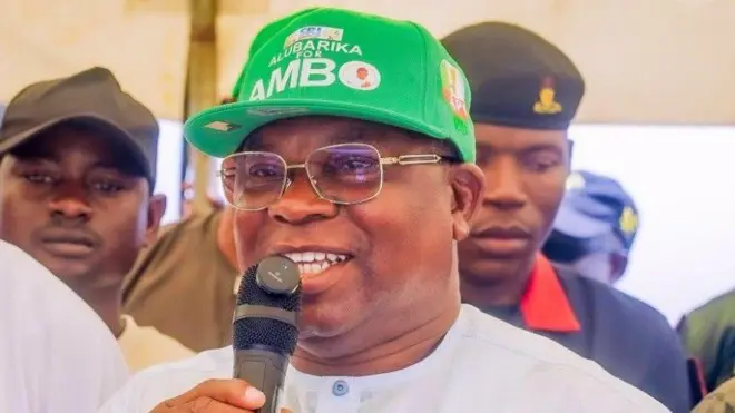 Asiwaju Munirudeen Bola Oyebamiji (AMBO), oludije sipo gomina Osun ninu ẹgbẹ APC. O de fila alawọ ewe, wọ aṣọ funfun. Awọn eeyan duro lẹyin rẹ bo ṣe n sọrọ si ẹrọ gbohungbohun to mu dani, bẹẹ lo n rẹrin-in musẹ pẹlu.