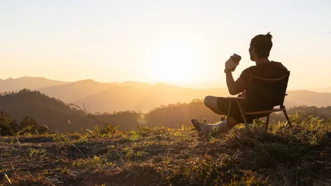 Чоловік сидить на стільці на траві та дивиться на гори, в руці в нього стаканчик з кавою
