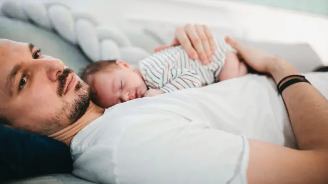 Un padre descansa en la cama con su bebé dormido sobre el pecho.