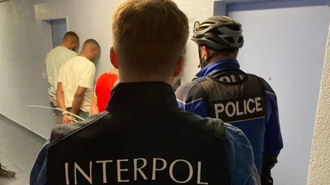 Two men wear interpol uniform stand wit three other men wey wey face wall