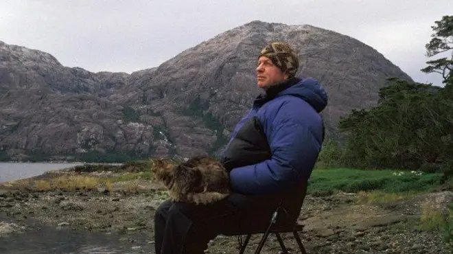 Bob Kull sentado en un silla con su gato en el regazo en la Patagonia chilena 