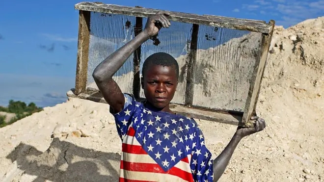 Un jeune garçon, en T-shirt aux couleurs du drapeau américain, se tient près d’une mine artisanale tenant un plateau de tamisage derrière ses épaules à Lumumbashi en RDC, décembre 2005.