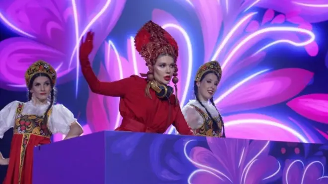 Geleneksel giysiler giyinmiş bir kadın şarkıcı ve iki kadın dansçı sahneden gösteri yapıyor. 