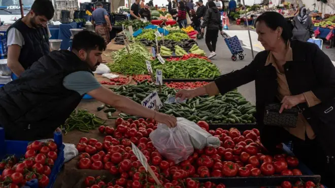 Bir pazarda domates salatalık biber ve diğer sebzelerin tezgahları yanyana uzanıyor. Bir kadın bir erkek satıcıdan domates alırken para uzatmış. Arka planda alışverişçiler ve satıcılar var.