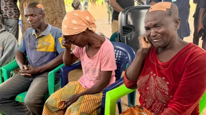 Three residents of Kurmin Wali village - two womrn and one man - sitted on plastic chairs. The women are crying, one of has plaster on her head. The man also has plaster on the back of his head as he looks forlorn into space.