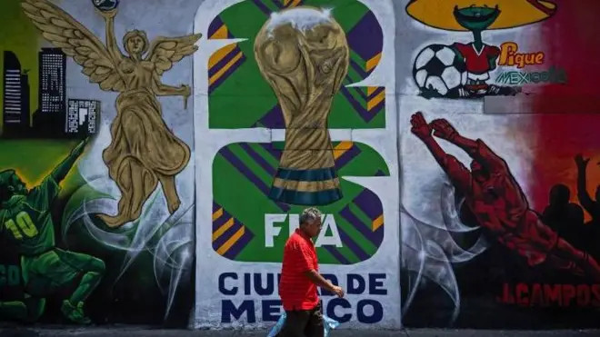 Un hombre con camisa roja camina frente a un mural en la Ciudad de México anuncia la llegada de la Copa Mundial de 2026, que se celebrará conjuntamente en México, Estados Unidos y Canadá.