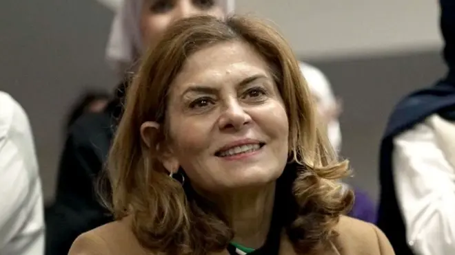 Close-up of Hind Kabawat, smiling. She has shoulder length brown hair and is wearing a smart, light brown coat.