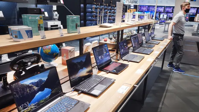 Магазин компьютерной техники. На заднем плане стоит продавец-консультант. На переднем - полка с включенными ноутбуками на продажу.