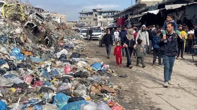 Palestinos desplazados caminan por un mercado junto a un vertedero de basura cubierto de bolsas de plástico. Un hombre pasa con un niño sobre los hombros, ambos mirando el vertedero. Detrás de ellos avanzan varios hombres, mujeres y niños. Al fondo se ven furgonetas blancas y edificios en ruinas.