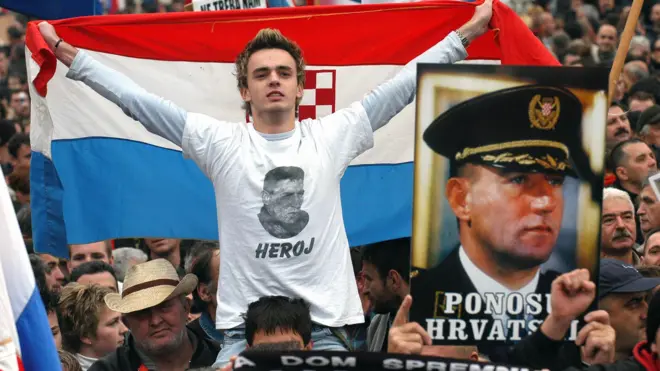 Молодой человек на плечах другого мужчины во время демонстрации. На нем белая футболка с черно-белым потретом Анте Готовины и надписью Heroj. За спиной у него хорватский флаг.