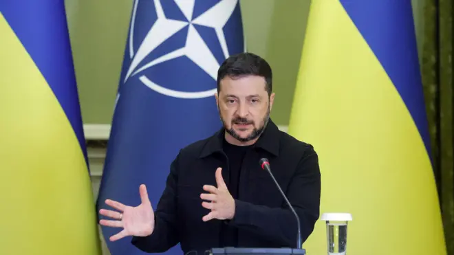 Perezida wa Ukraine Volodymyr Zelensky, yambaye imyenda y'ibara ry'igikara akunze kwambara, ahagaze akora ikimenyetso n'ibiganza, avugira mu ndangururamajwi, mu kiganiro n'abanyamakuru mu murwa mukuru Kyiv. Inyuma ye hari ibendera rya Ukraine.