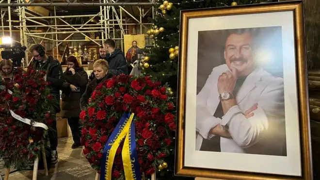 Портрет усміхненого Степана Гіги у білому костюмі. Поряд – вінки з червоних гвоздик, один з яких зі стрічкою із написом "від президента України"