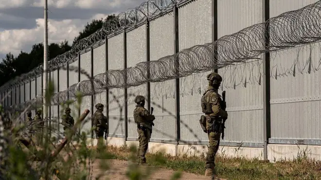 Soldados fronterizos armados, montan guardia frente a una larga y alta valla metálica con concertinas de alambres de púas