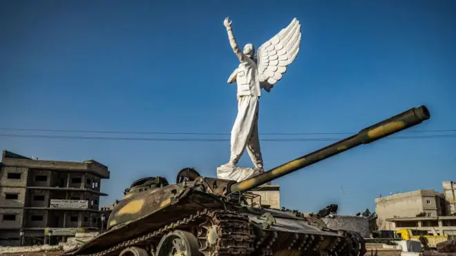 صورة تظهر تمثالاً أمامه دبابة مدمرة وسط ميدان في مدينة كوباني أو عين العرب شمالي سوريا.