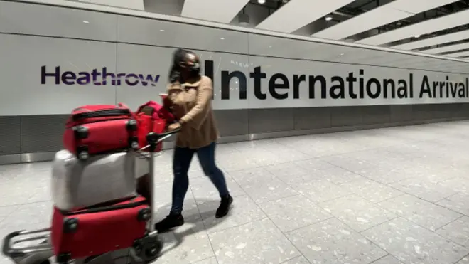 One passenger dey waka through di arrivals area of Heathrow Airport for London, England. She dey pull suitcase.