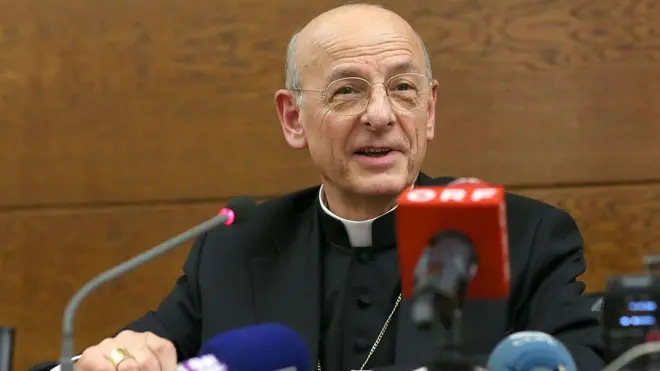 Fernando Ocáriz Braña sorrindo ao falar no microfone; ele está sentado em uma sala, diante de vários microfones