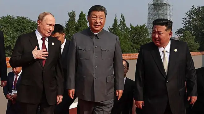 Vladimir Putin, Xi Jinping e Kim Jong Un, lado a lado, todos de terno e outras pessoas de terno atrás deles. Putin está com a mão direita no peito.